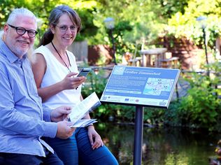 Zwei Personen mit Handy und Flyer vor einem Info-Schild am Wasser. Das Schild zeigt eine Karte mit Stationsmarkierungen und weitere Infos.