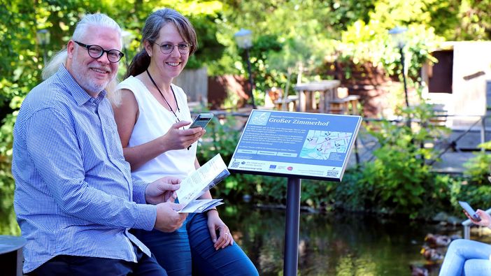 Zwei Personen mit Handy und Flyer vor einem Info-Schild am Wasser. Das Schild zeigt eine Karte mit Stationsmarkierungen und weitere Infos.