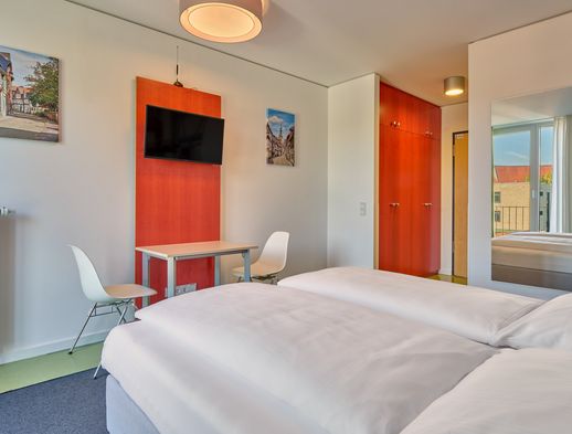 Modernes Doppelzimmer mit zwei Betten, TV, Schreibtisch, großem Spiegel und hellem Tageslicht.