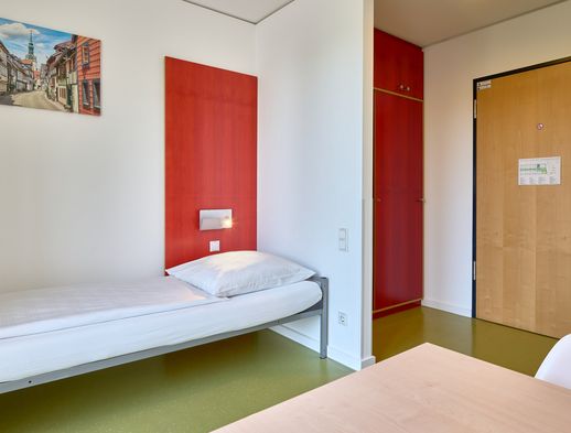 Einzelzimmer mit schlichtem Bett, rotem Wandelement, Holztür und Wandbild der Altstadt Wolfenbüttels.