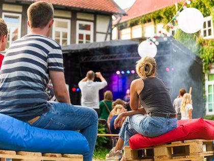 Rückenansicht von Personen in T-Shirts, 3 im Vordergrund sitzen auf Palettenmöbeln, im Hintergrund eine Bühne, dekorative Lampions