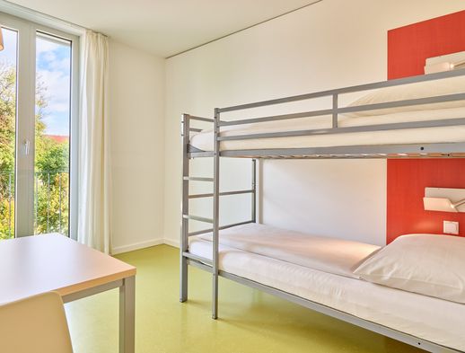 Helles Zimmer mit Etagenbett, großem Fenster, grünem Boden und schlichtem Tisch mit Stühlen.