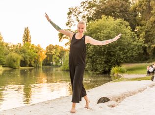 Eine lachende Frau balanciert barfuß auf einem Balken am Wasser